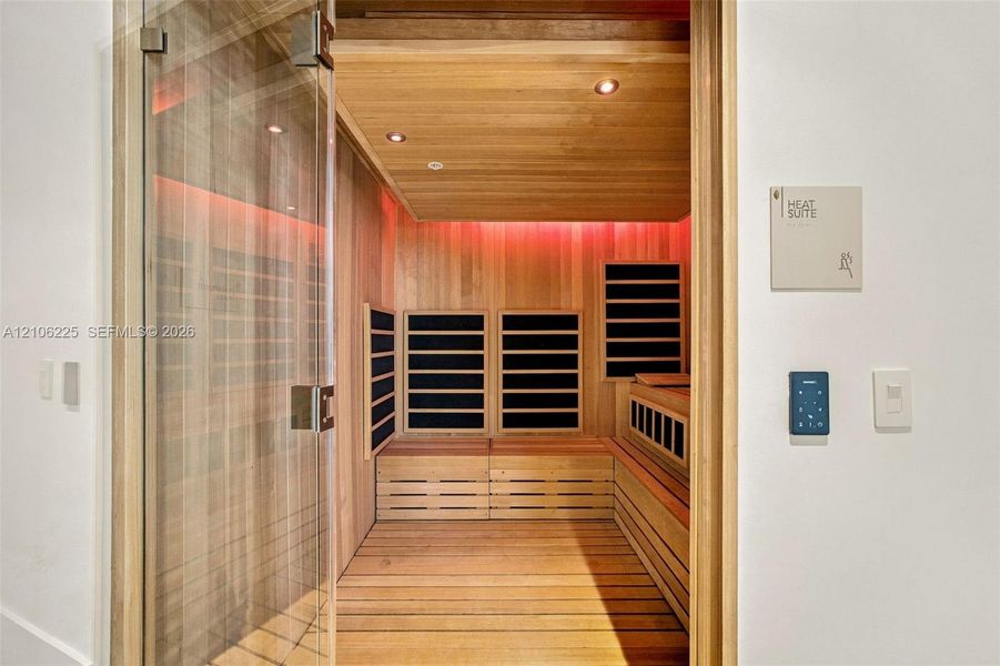 Sauna room featuring light wood-paneled walls and flooring, integrated heating elements, recessed lighting, and a glass door with a polished metallic handle Sauna room featuring light wood-paneled walls and flooring, integrated heating elements, recessed lighting, and a glass door with a polished metallic handle