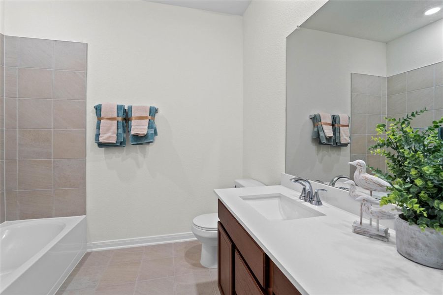 The secondary bathroom showcases tile flooring, a bath/shower combo with tile surround, a vanity with knee space and a stunning light countertop, a stylish mirror, sleek fixtures, and contemporary finishes throughout.