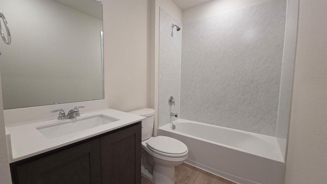 Bathroom featuring a dark wood-finish vanity with a white countertop and integrated sink, a large wall-mounted mirror, a standard toilet, and a white bathtub with a textured surround Bathroom featuring a dark wood-finish vanity with a white countertop and integrated sink, a large wall-mounted mirror, a standard toilet, and a white bathtub with a textured surround