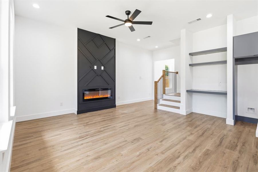 Spacious room with wood-finish flooring, a contemporary black accent wall featuring an electric fireplace, built-in shelving, and a modern ceiling fan Spacious room with wood-finish flooring, a contemporary black accent wall featuring an electric fireplace, built-in shelving, and a modern ceiling fan