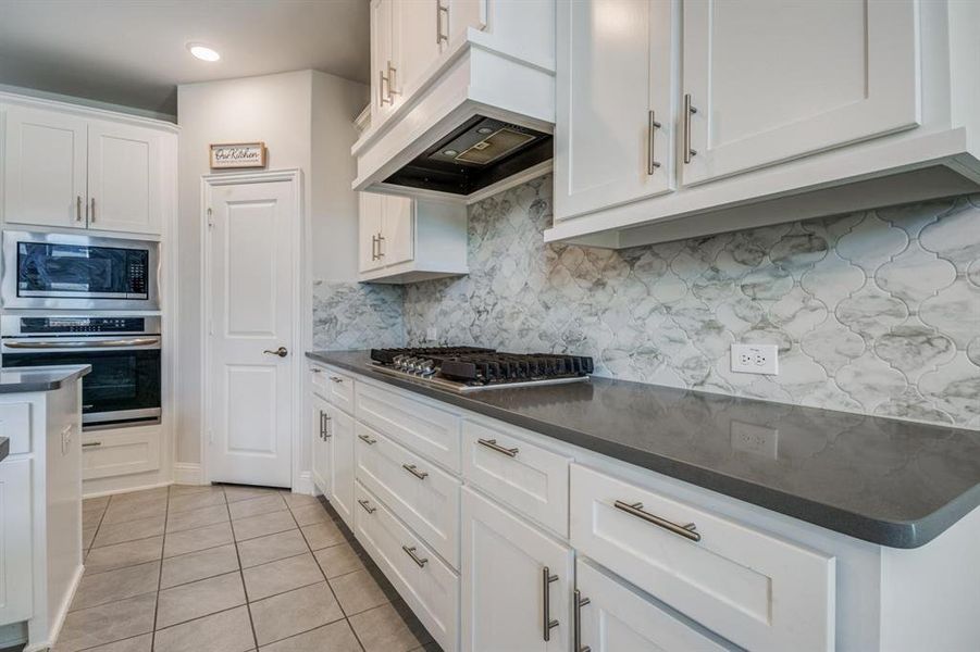 Kitchen with stainless steel appliances, backsplash, light tile patterned flooring, custom exhaust hood, and white cabinetry