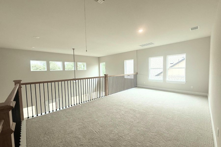 Spacious loft area featuring light gray carpeting, multiple windows, recessed lighting, and a wood-finish railing with black metal balusters Spacious loft area featuring light gray carpeting, multiple windows, recessed lighting, and a wood-finish railing with black metal balusters