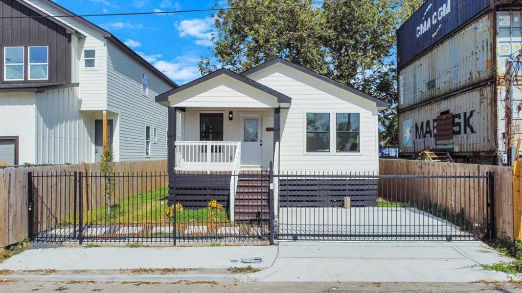 This is a compact, single-story home featuring a fresh, modern design with a cozy front porch, highlighted by a secure, black metal fence enclosing a small yard. The property is situated next to a large, two-story house and in proximity to industrial-style structures.