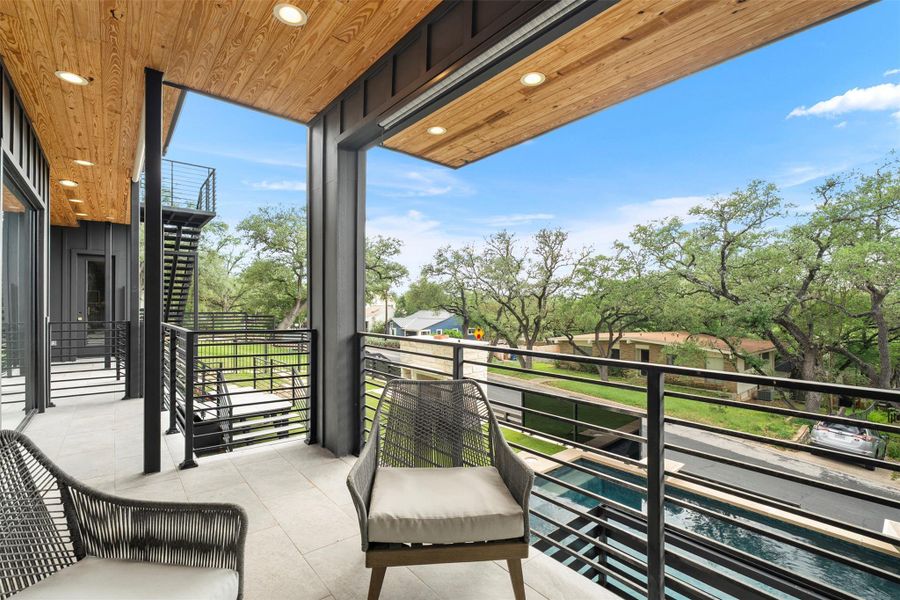 Expansive outdoor terrace featuring a wood-paneled ceiling with recessed lighting, tile flooring, and metal railings overlooking mature trees and a swimming pool