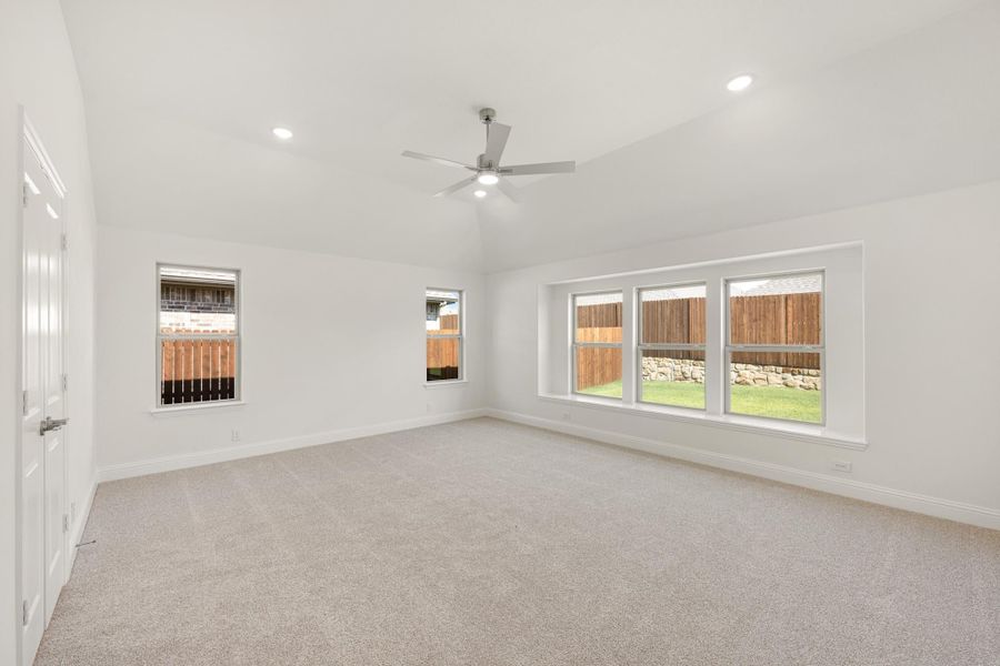 Empty bedroom with carpet, ceiling fan, vaulted ceiling, and multiple windows overlooking backyard