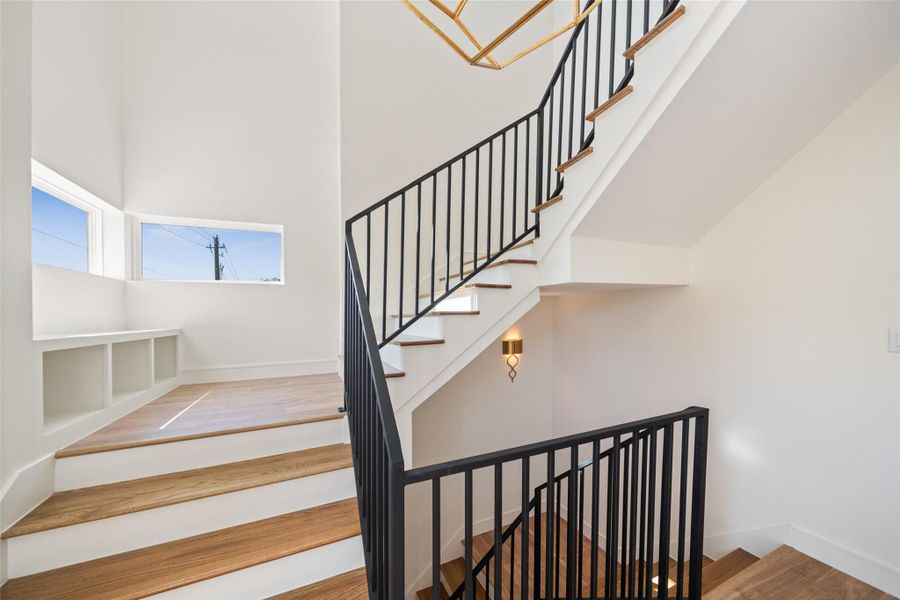 Modern staircase with sleek black railings and wooden steps. The space is bright and airy, featuring large windows that allow natural light to fill the area. A stylish wall sconce adds an elegant touch.
