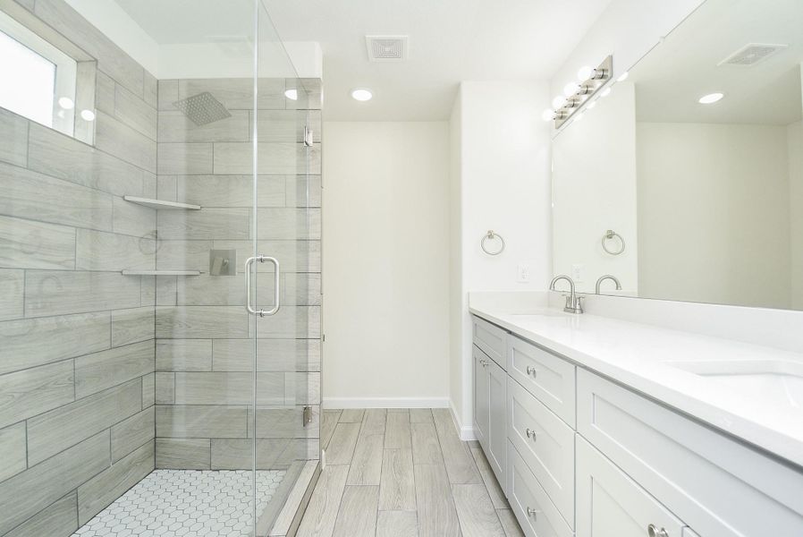 This is a modern bathroom featuring a large walk-in shower with gray wood-look tile, a clear glass door, and a white hexagon-tiled floor. There's a spacious double vanity with white countertops, ample storage, and a well-lit mirror above. The space has a clean, minimalist aesthetic. This is a modern bathroom featuring a large walk-in shower with gray wood-look tile, a clear glass door, and a white hexagon-tiled floor. There's a spacious double vanity with white countertops, ample storage, and a well-lit mirror above. The space has a clean, minimalist aesthetic.