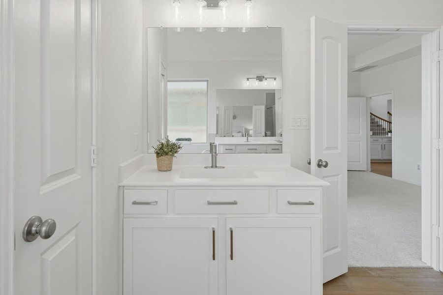Bright and modern bathroom with a sleek white vanity, large mirror, and stylish lighting. The room features a clean, minimal design with access to an adjoining space through open doors, leading to a well-lit interior with carpeted flooring.