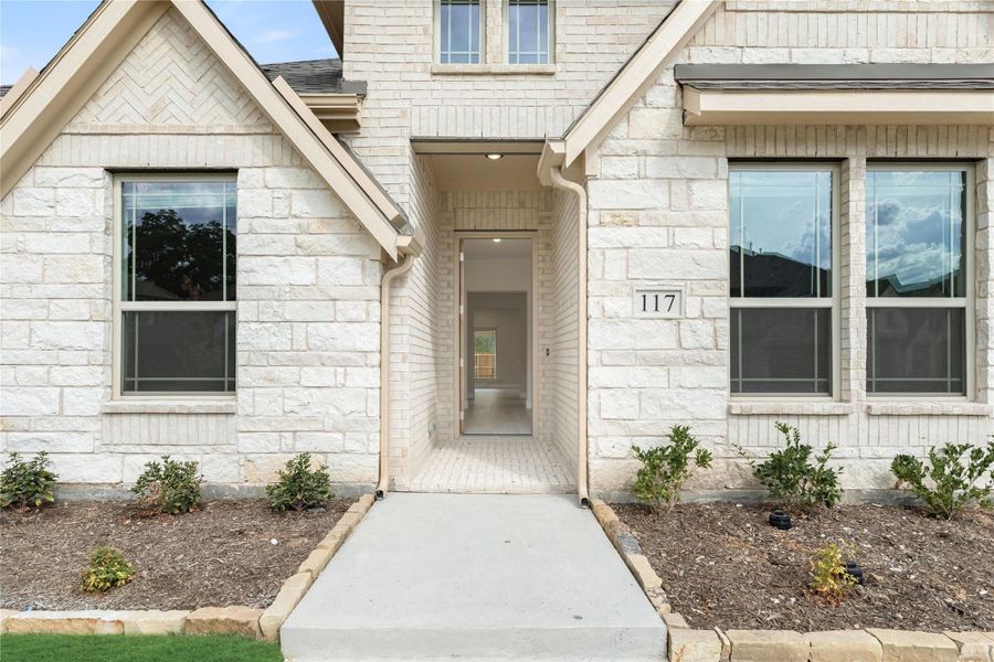 Front entry of a light stone brick home with covered doorway, concrete walkway, and address number 117.