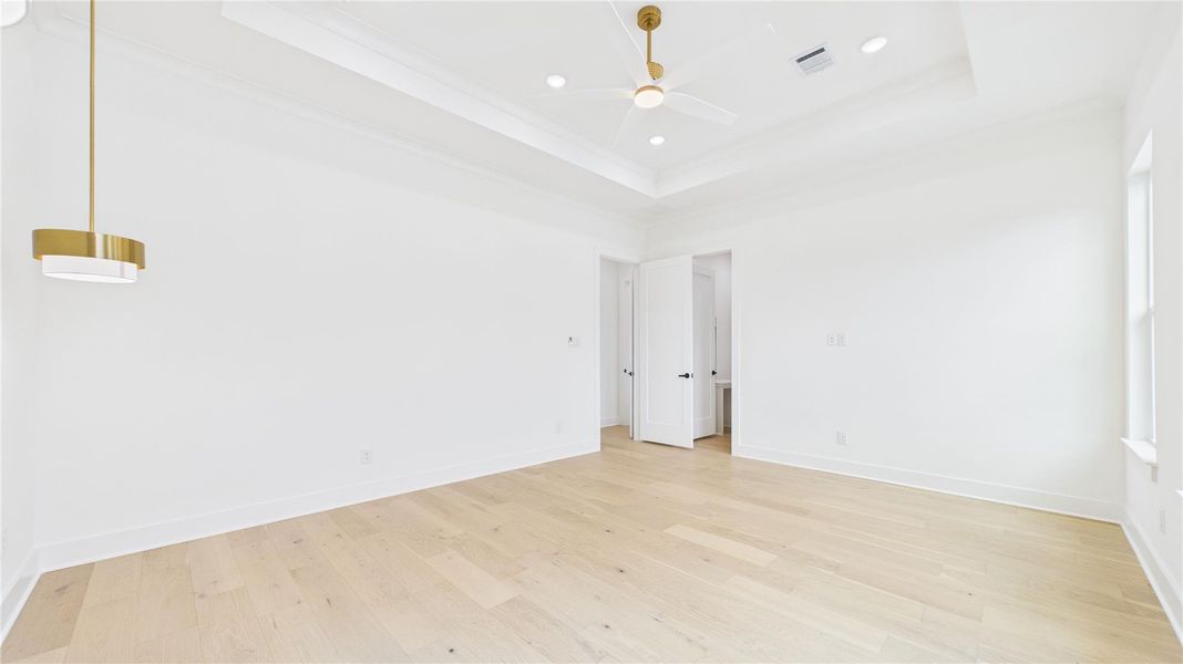 Huge primary bedroom filled with natural  light, a continuation of hardwood floors and white walls. Features a modern ceiling fan, recessed lighting, and stylish pendant lights for reading in bed. The room has a tray ceiling.