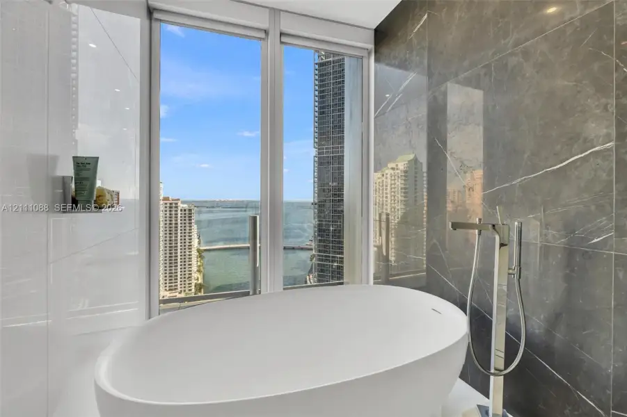 Freestanding soaking tub with polished chrome floor-mounted faucet, large-format grey stone wall tile, and expansive floor-to-ceiling windows Freestanding soaking tub with polished chrome floor-mounted faucet, large-format grey stone wall tile, and expansive floor-to-ceiling windows