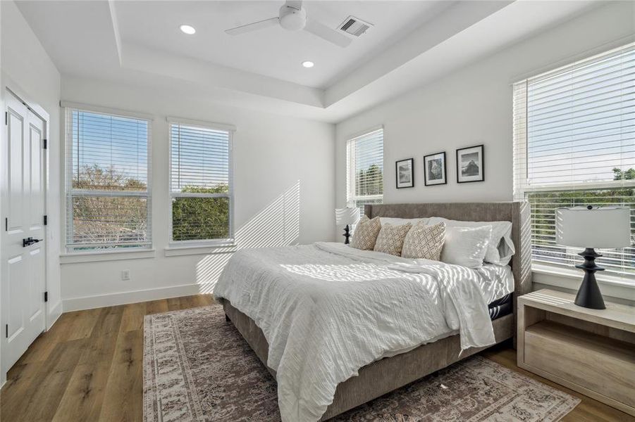 Another view of the primary bedroom featuring tray ceilings and and abundance of windows for natural light.