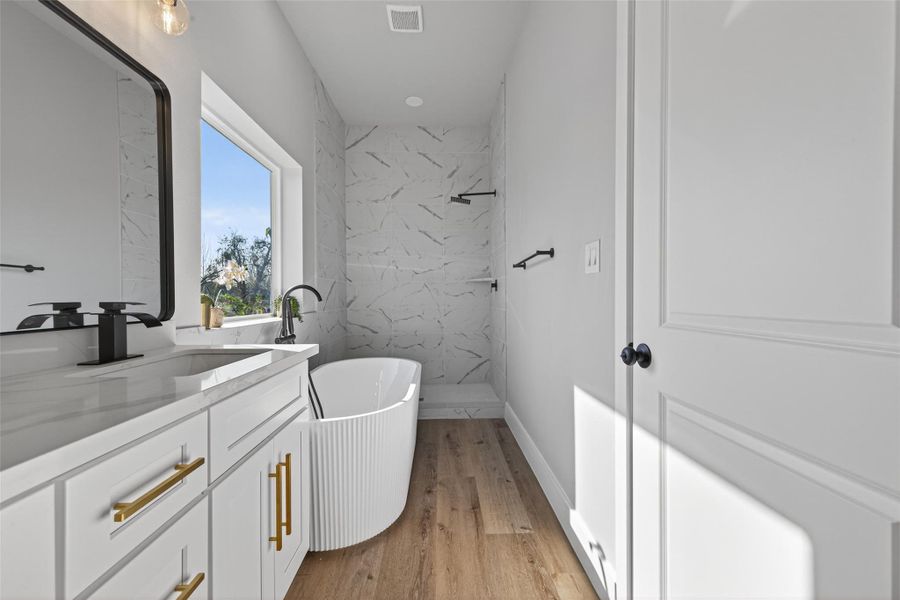 Luxe freestanding soaking tub paired with a floor-mounted matte black filler and a frameless walk-in shower featuring an oversized matte black rainfall head, all set against full-height marble-look tile walls. Natural light, recessed lighting, and water-efficient fixtures create a serene, spa-inspired retreat designed for both indulgence and sustainability.