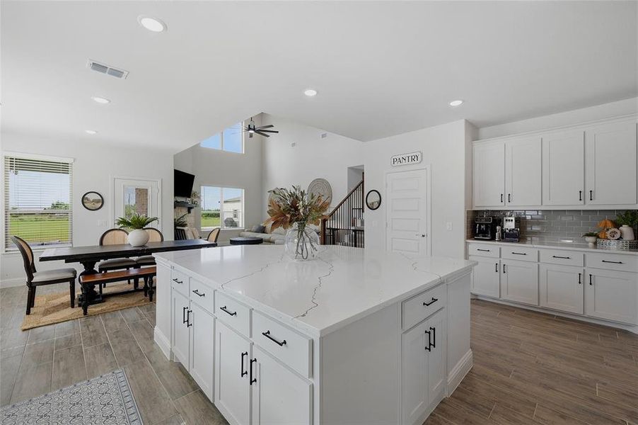 Spacious kitchen featuring a large central island with white cabinetry and a light-toned countertop, complemented by a full wall of white upper and lower cabinetry with dark hardware and a gray subway tile backsplash