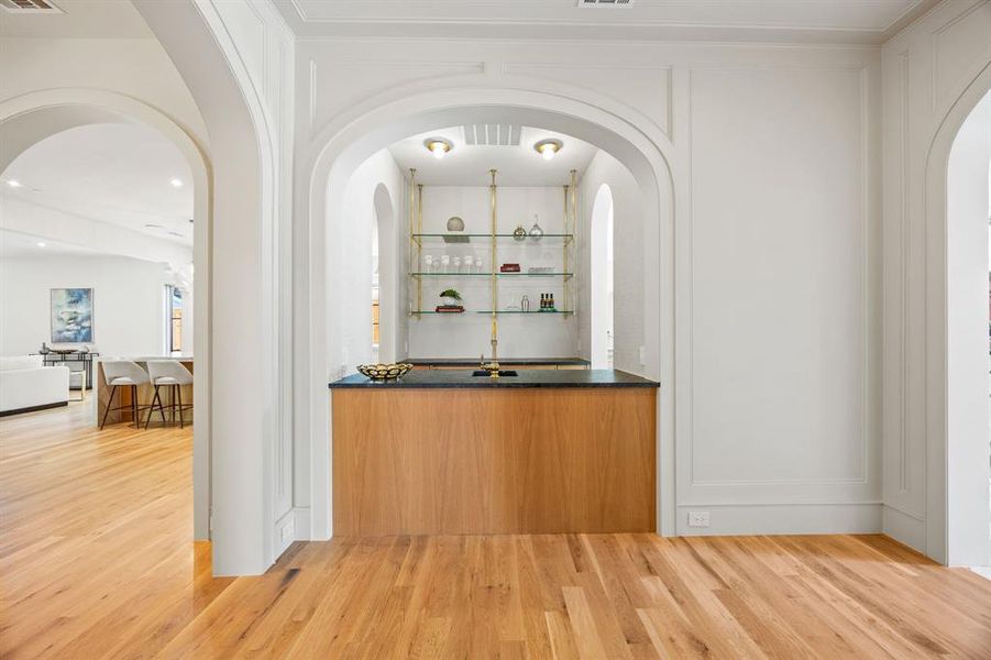 Built-in bar featuring a dark countertop, light wood-finish cabinetry, and brass-toned shelving with glass inserts Built-in bar featuring a dark countertop, light wood-finish cabinetry, and brass-toned shelving with glass inserts