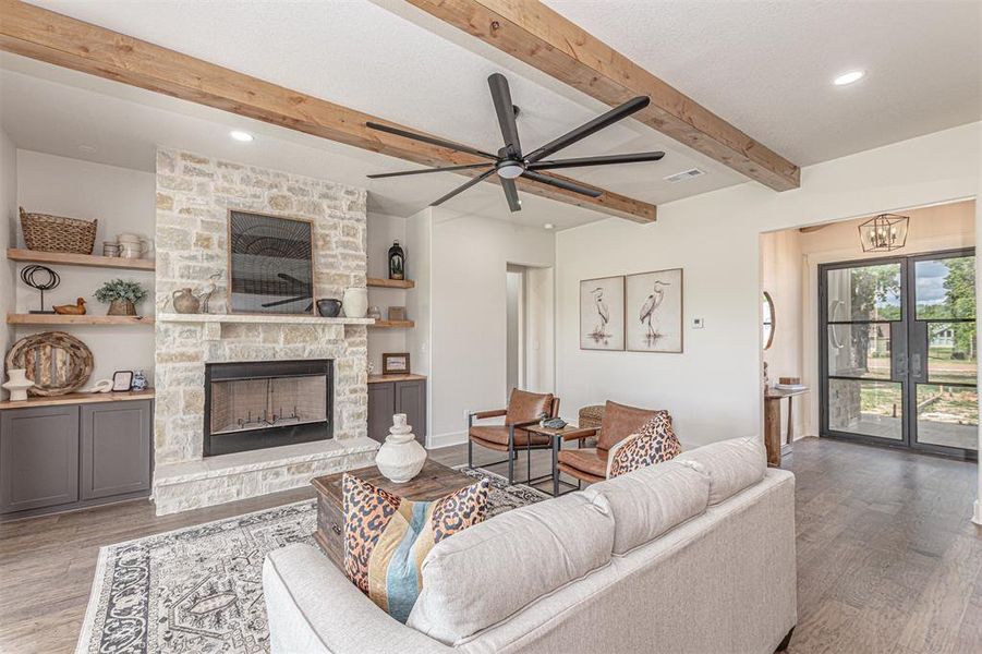 Living area featuring a ceiling fan, wood finished floors, a stone fireplace, beam ceiling, and suspended lighting