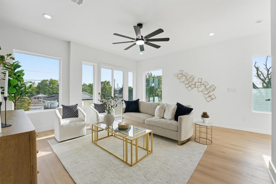 Bright, modern living room featuring light wood-look flooring, oversized windows, recessed lighting, and a contemporary ceiling fan, creating an open, sun-filled space ideal for relaxing or entertaining.