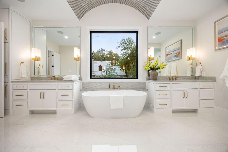 A perfectly balanced view of the primary bathroom showcases symmetry and sophistication, with dual vanities flanking a central soaking tub. Large mirrors and soft lighting enhance the bright, airy feel. The highlight of this space is the centered freestanding tub beneath the window, creating a visually striking focal point that embodies both relaxation and timeless design.