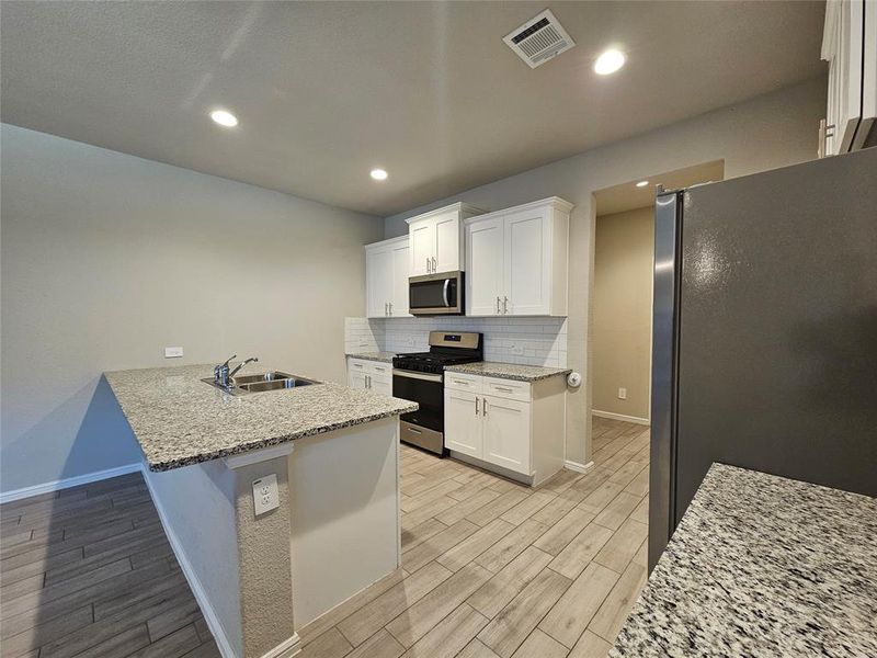 Kitchen with white cabinetry, open floor plan, stainless steel appliances, recess lights, granite counters, backsplash and wood looking tiled floors.