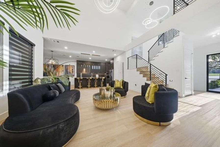 Open-concept main living area with high ceilings, blonde wood-finish flooring, a modern horizontal bar staircase, built-in bar with pendant lighting, and large windows with horizontal blinds