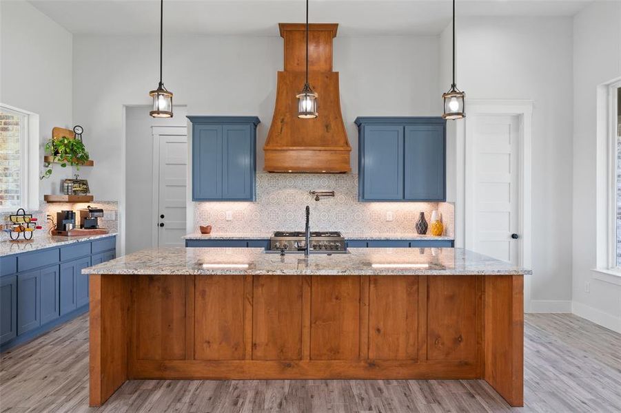 Spacious kitchen featuring a large wood-finish island, gray-flecked countertops, blue cabinetry, a decorative wood-finish range hood, and pendant lighting