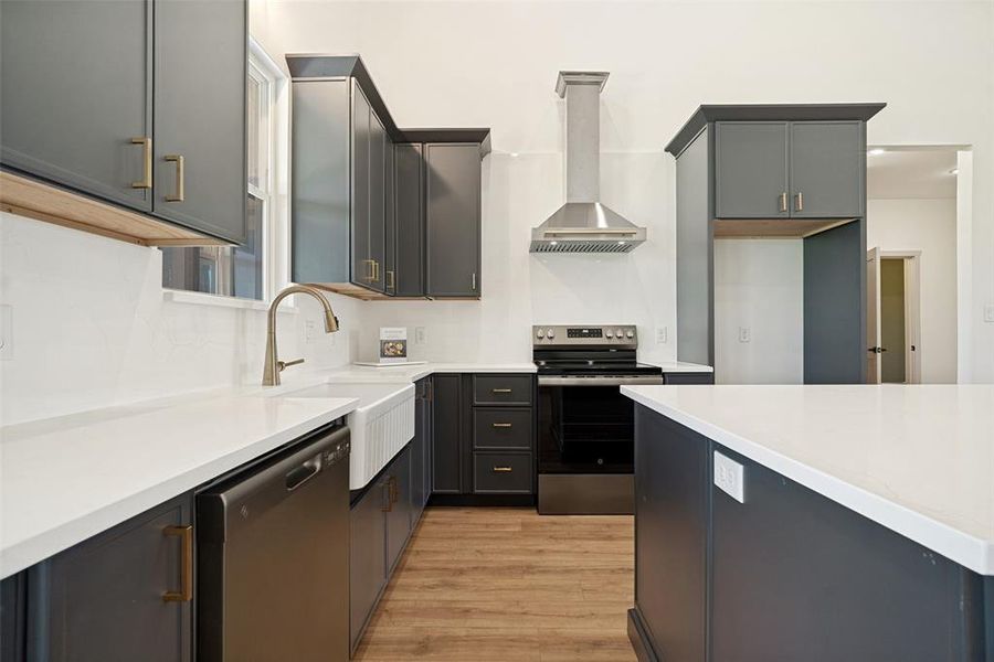 Modern kitchen featuring white countertops, dark gray cabinetry with brass hardware, wood-finish flooring, a stainless steel range hood, and a white farmhouse sink
