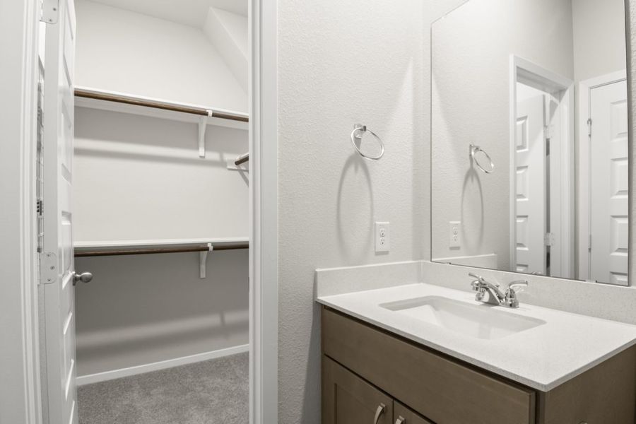 Image of a primary bathroom with brown cabinets, a white vanity, a mirror and a walk-in closet