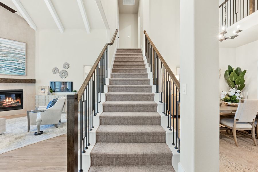 Carpeted staircase with wood handrail and black iron balusters, flanked by living room with fireplace and dining area Carpeted staircase with wood handrail and black iron balusters, flanked by living room with fireplace and dining area