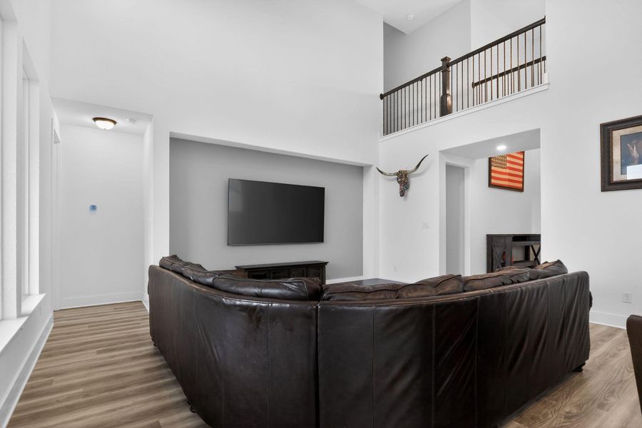 Spacious living area featuring high ceilings, wood-finish flooring, a contemporary wall-mounted television, and an upper-level balcony with dark wood railing Spacious living area featuring high ceilings, wood-finish flooring, a contemporary wall-mounted television, and an upper-level balcony with dark wood railing