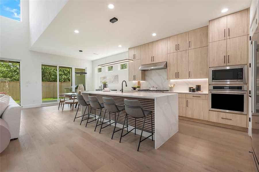 Kitchen featuring light brown cabinets, light wood-type flooring, an island with sink, appliances with stainless steel finishes, and a kitchen bar