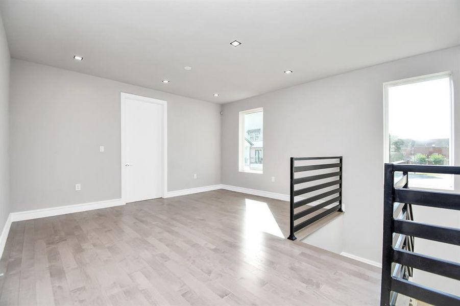 This is a bright, spacious room with modern gray flooring, large windows providing natural light, and a contemporary black railing. The room features recessed lighting and has a clean, minimalist look.