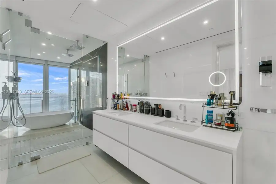 Floating double vanity with dual undermount sinks, expansive LED-backlit vanity mirror, glass-enclosed wet room featuring a freestanding soaking tub, and floor-to-ceiling windows Floating double vanity with dual undermount sinks, expansive LED-backlit vanity mirror, glass-enclosed wet room featuring a freestanding soaking tub, and floor-to-ceiling windows
