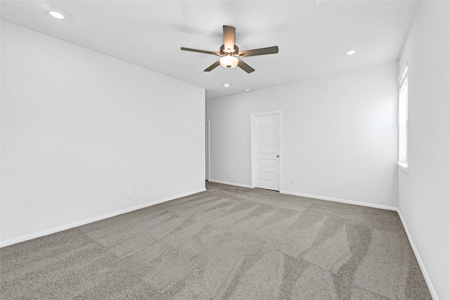 This photo showcases a spacious, carpeted bedroom with neutral walls, recessed lighting, and a ceiling fan. It features a window for natural light and a door leading to an adjacent room or closet.Please note: The image is not the actual home; it is a home with the same floor plan in the neighborhood.