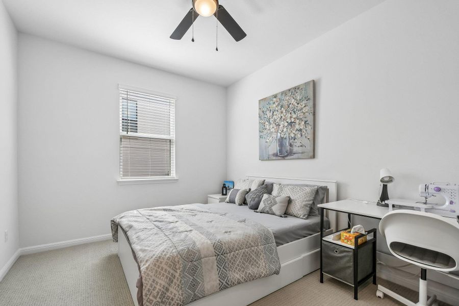 A well-lit bedroom features a ceiling fan, plush carpet, and a window for natural light.