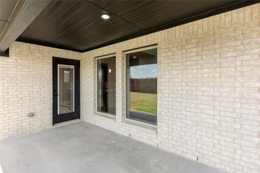 Covered back patio with concrete floor, light brick exterior, black door, and large windows overlooking a grassy yard Covered back patio with concrete floor, light brick exterior, black door, and large windows overlooking a grassy yard