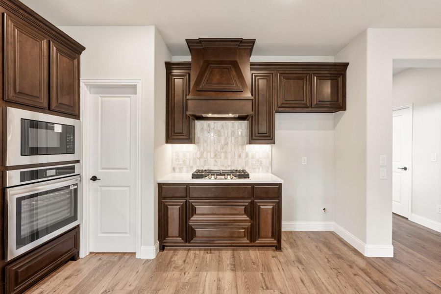 Kitchen with dark wood cabinets, wood range hood, gas cooktop, double wall oven, and light hardwood floors