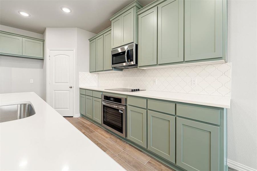 Kitchen featuring green cabinets, decorative backsplash, appliances with stainless steel finishes, wood finish floors, and light stone countertops Kitchen featuring green cabinets, decorative backsplash, appliances with stainless steel finishes, wood finish floors, and light stone countertops