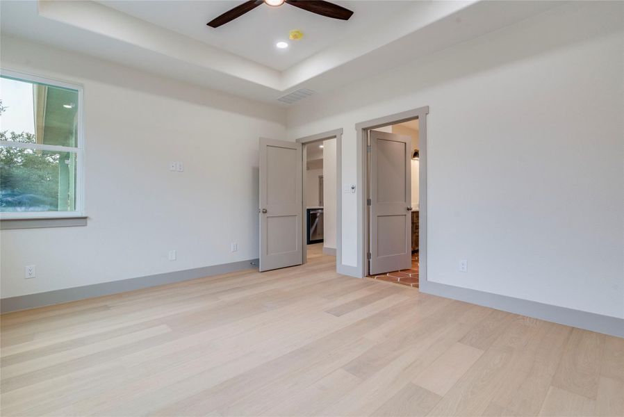Unfurnished bedroom featuring a raised ceiling, light wood-type flooring, ceiling fan, ensuite bath, and recessed lighting