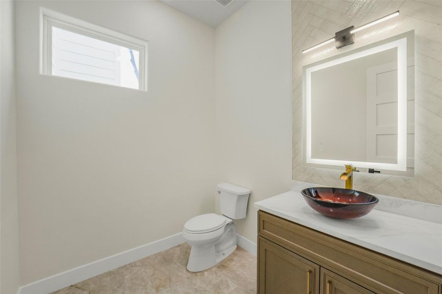 This bathroom features a modern design with a wall-mounted lighted mirror, elegant vessel sink, and gold faucet on a marble countertop. It has a minimalist aesthetic with neutral tones and a small window for natural light.