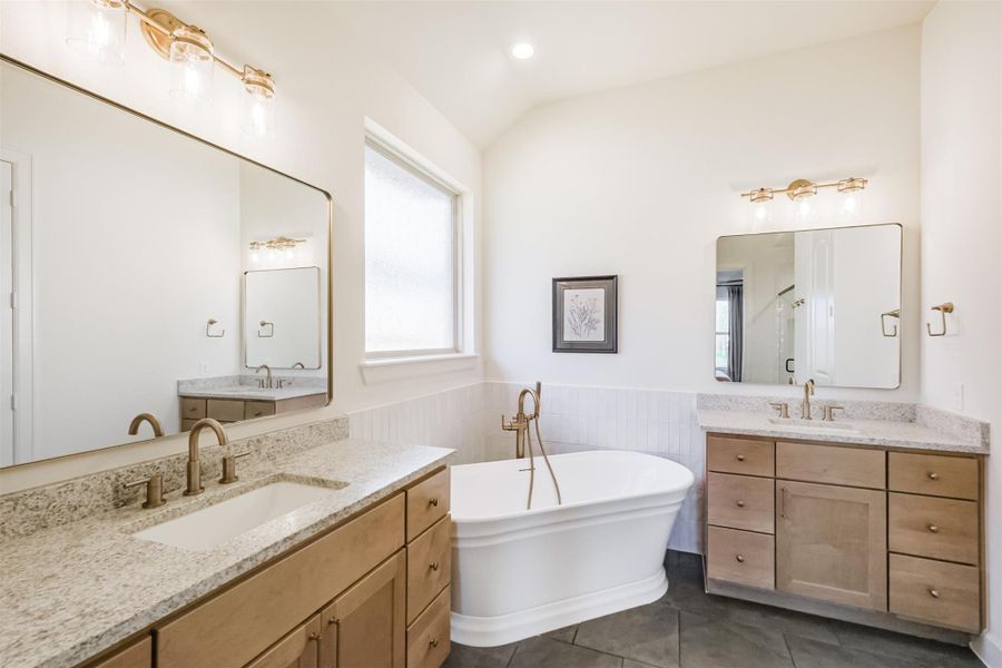 View of the luxurious primary bathroom showcasing light wood vanity, dual sinks, granite counters, and large mirrors.