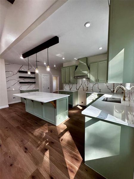 Spacious kitchen featuring a large central island with a white countertop, light green cabinetry, wood-finish flooring, a decorative range hood, and a white backsplash with dark veining