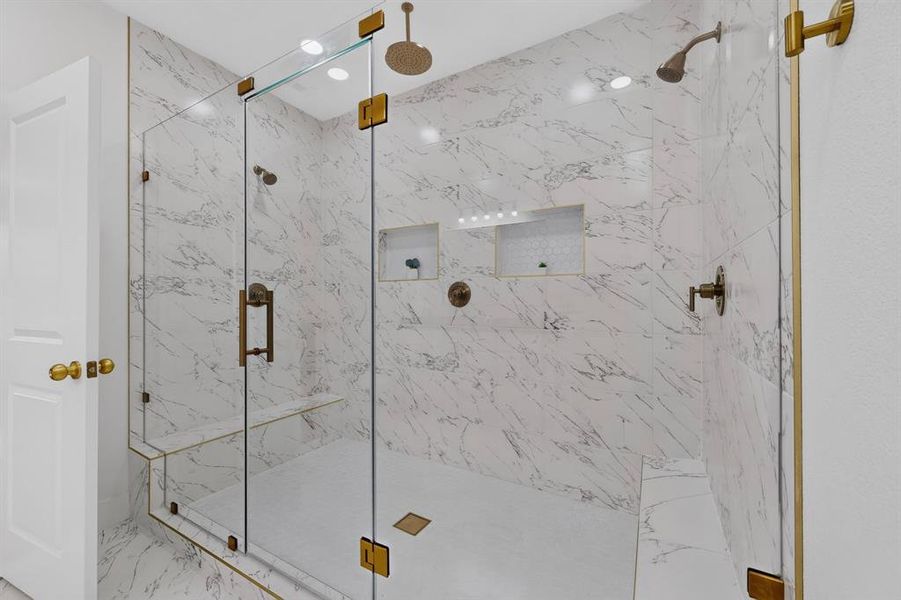Spacious glass-enclosed shower featuring large format marble-look tile, dual shower heads, integrated bench seating, recessed niches, and brass-finish hardware