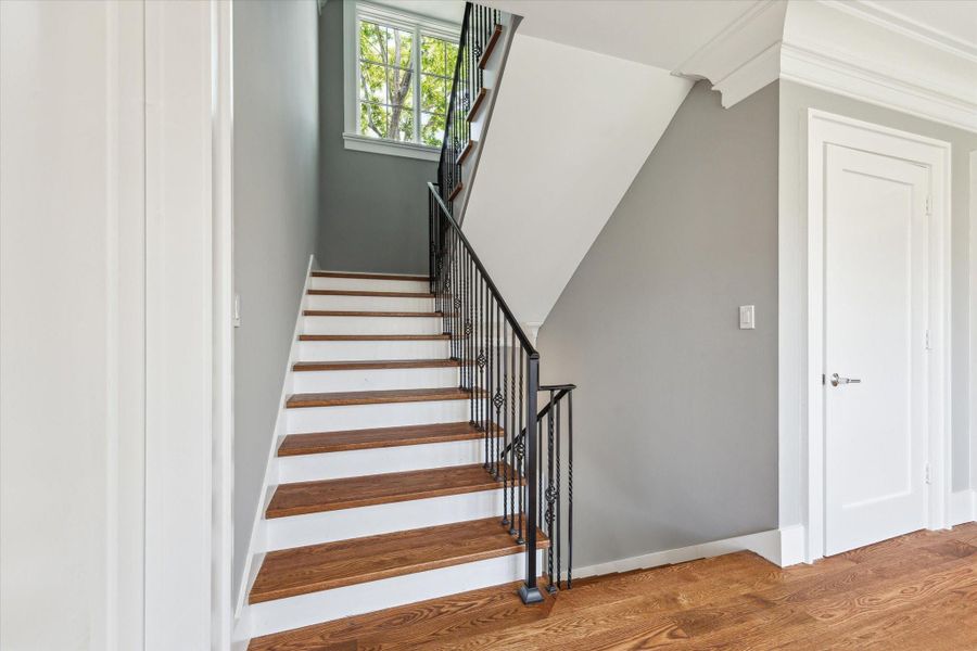 A bright and welcoming staircase with wooden steps and a black metal railing leads to the upper floor. The door to the right is the door for the elevator capable closet.  The space features light gray walls, white trim, and a window providing natural light.