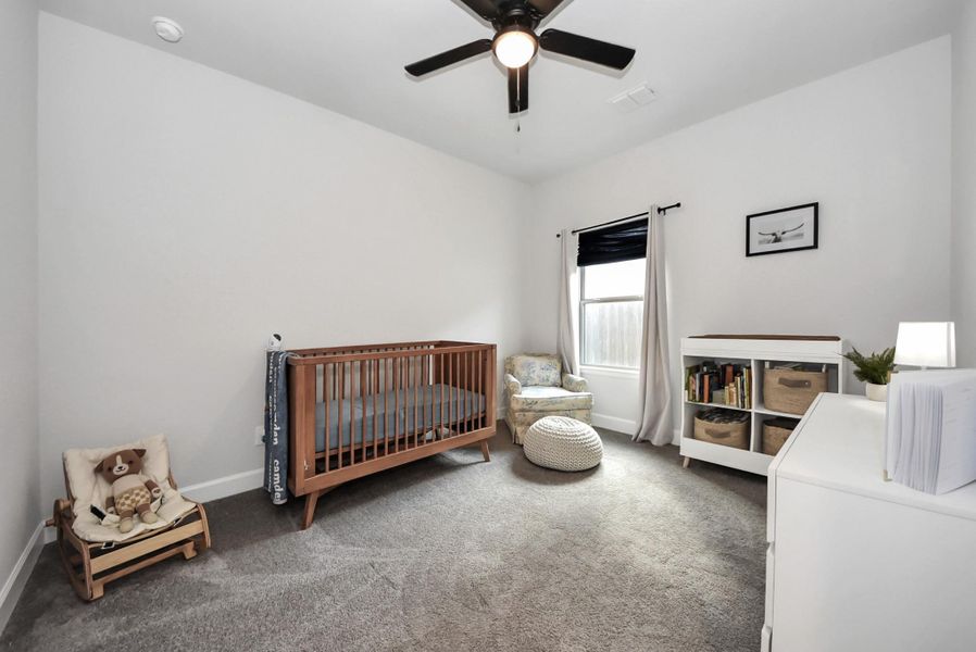 This charming nursery features soft neutral walls, a ceiling fan, and natural light streaming through a curtained window. Thoughtfully arranged with a wooden crib, cushioned chair, soft pouf, and organized shelving, the room provides a calm and welcoming space for a little one.