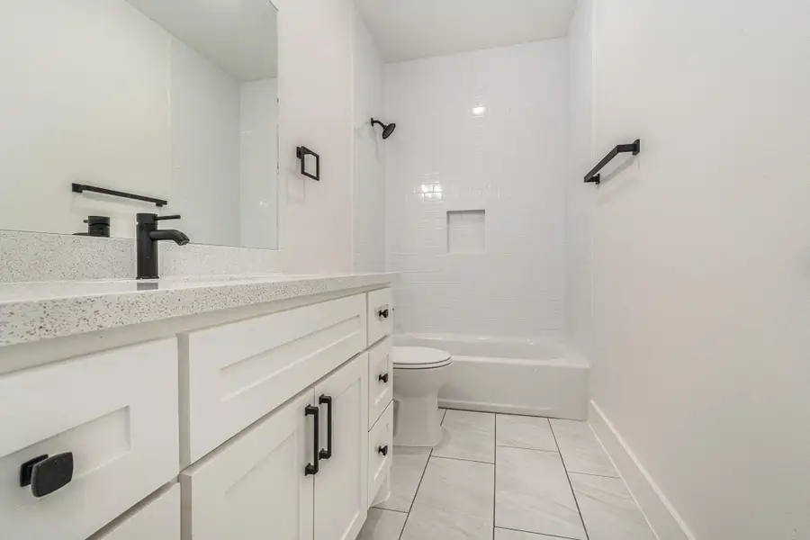 This bathroom features a modern, clean design with white cabinetry and countertops, black fixtures, and a tiled shower-tub combination. The room is bright and spacious, offering a sleek and functional space.