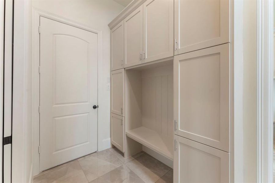 This is a clean and modern mudroom with built-in storage cabinets and a bench, featuring neutral colors and tile flooring. Ideal for organizing outerwear and shoes. The door leads to your spacious 3 car garage.