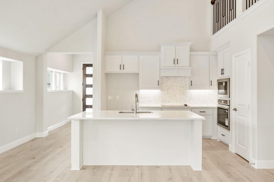 White kitchen with large island, white cabinets, marble backsplash, double wall ovens, and light hardwood floors