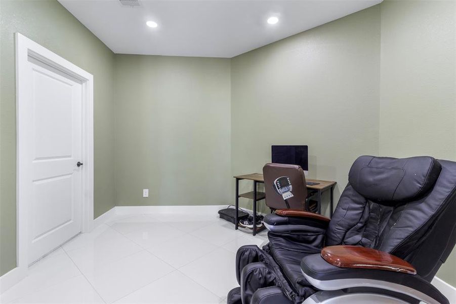 This photo shows a cozy room with light green walls and glossy white tile flooring. It features a comfortable black massage chair and a small desk with a computer, creating a perfect space for relaxation or work. The room is well-lit with recessed lighting and has a simple, modern door.