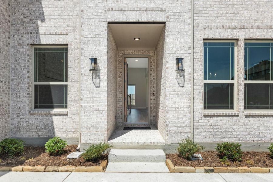 Home entrance with white brick exterior, recessed entry, two wall sconces, and concrete steps with landscaping Home entrance with white brick exterior, recessed entry, two wall sconces, and concrete steps with landscaping