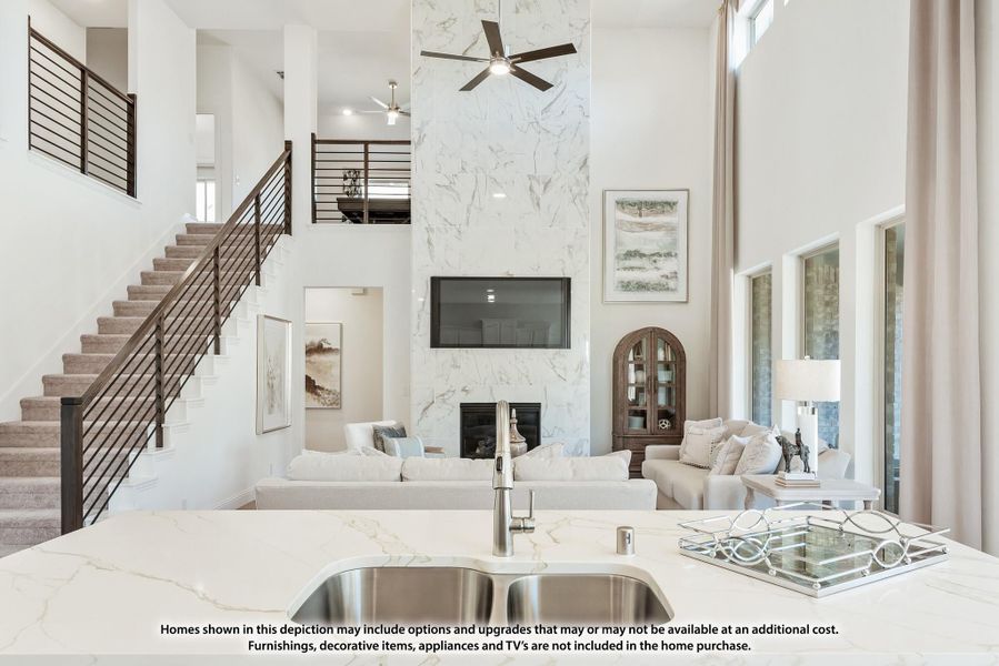 Open-concept living area with two-story marble fireplace, kitchen island sink, and staircase with metal railing Open-concept living area with two-story marble fireplace, kitchen island sink, and staircase with metal railing